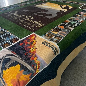 May include: A quilted blanket featuring the text "Great Smoky Mountains National Park." The quilt has a green border and includes images of the park's scenery. The quilt is made of various fabrics and colors, including brown, yellow, and blue.