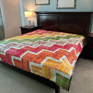 May include: A colorful chevron patterned quilt in shades of green, yellow, orange, pink, and white is displayed on a bed with a dark wood headboard. The quilt is made with a variety of fabrics and has a white border.