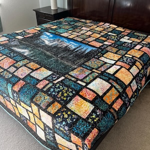 May include: A colorful quilt featuring a central landscape scene of mountains and a lake. The quilt is bordered by a patchwork design in various colors including teal, orange, and black. The quilt is displayed on a dark wood bed frame.