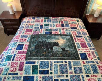 May include: A colorful quilt with a patchwork design in shades of blue, pink, and purple. The quilt features a central panel depicting a bear family in a forest setting. The quilt is placed on a bed with dark wood furniture.
