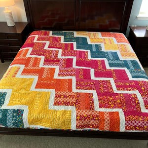 May include: A colorful patchwork quilt with a chevron pattern in shades of teal, yellow, orange, and red. The quilt is on a bed with a dark wood headboard.