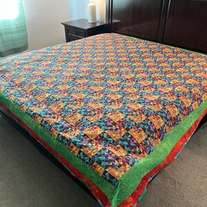 May include: A colorful quilt with a geometric pattern of squares and triangles in shades of orange, blue, green, and purple. The quilt is on a bed with a dark wooden frame. The edges are trimmed with green and red fabric.