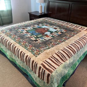 May include: A large, colorful quilt with a central square featuring a bear in a forest scene. The quilt has a patterned border with brown and cream stripes, and green and blue accents. The quilt is on a bed with a dark brown headboard.