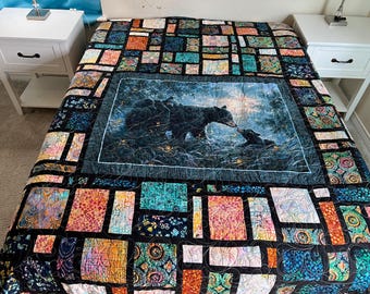 May include: A colorful quilt featuring a central image of a mother bear and cub in a forest setting. The quilt has a patchwork design with various rectangular blocks in shades of blue, orange, and green, creating a vibrant and cozy aesthetic.