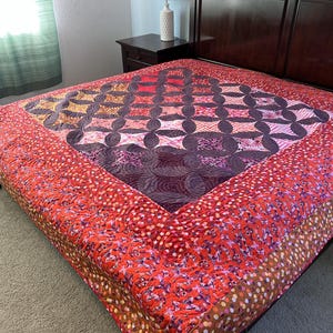 May include: A handmade quilt with a vibrant design. The center features a geometric pattern in shades of purple, orange, and brown. The border is a red floral print. The quilt is displayed on a bed with a dark wood headboard.