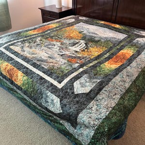 May include: A quilted bedspread featuring a landscape design with a waterfall, trees, and mountains. The quilt has a central square image with a white border, surrounded by patterned sections in shades of green, orange, and gray.