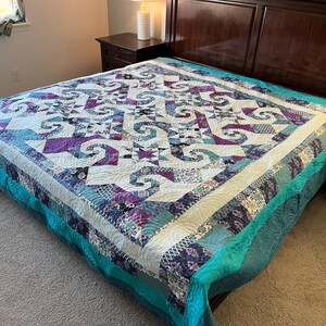 May include: A handmade quilt with a complex geometric pattern in shades of purple, teal, and white. The quilt is spread across a bed, showcasing its intricate design and craftsmanship. The quilt's border is a solid teal color.