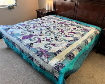 May include: A handmade quilt with a complex geometric pattern in shades of purple, teal, and white. The quilt is spread across a bed, showcasing its intricate design and craftsmanship. The quilt's border is a solid teal color.