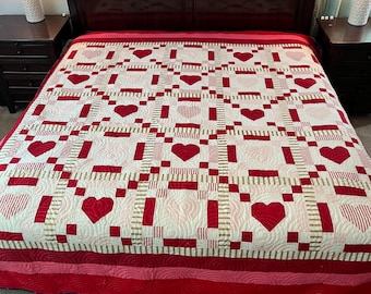 May include: A red and white quilt with a heart pattern. The quilt features large red hearts and a patchwork design with red and gold accents. The quilt is on a bed with a dark wood headboard and nightstands.