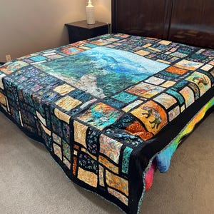 May include: A large patchwork quilt featuring a central rectangular panel depicting a blue mountain landscape. The surrounding border consists of various smaller fabric squares in shades of orange, teal, blue, and black, creating a complex grid pattern.