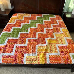 May include: A colorful quilt with a chevron pattern in shades of orange, yellow, green, and white. The quilt is made of patchwork fabric and is displayed on a bed.