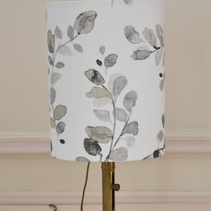 Peut inclure: Lampe de table en laiton avec abat-jour cylindrique. L'abat-jour est blanc avec un motif répété de feuilles et de tiges grises et beiges de style aquarelle. La lampe a une base ronde et un cordon.