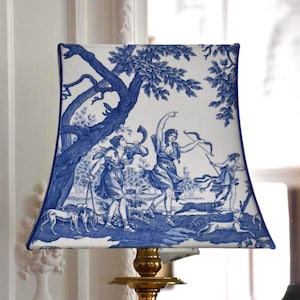 Puede incluir: Pantalla de lámpara cuadrada con una escena pastoral azul y blanca. El diseño presenta figuras, árboles y perros, con un borde azul oscuro. La pantalla está montada sobre una base de latón.