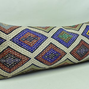 May include: A long, rectangular cushion with a white background and a geometric pattern in shades of blue, red, green, and yellow. The pattern is made up of squares and diamonds.