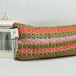 May include: A long, rectangular cushion with a colourful geometric pattern. The cushion is made of woven fabric and has a green, pink, and orange colour scheme. The cushion is sitting on a white surface.