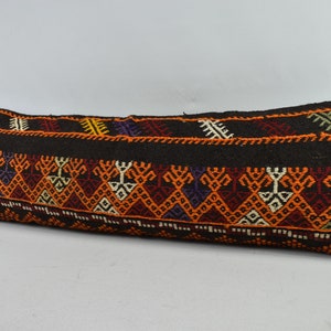 May include: A long, rectangular pillow with a black background and colorful geometric patterns in shades of orange, red, purple, and green. The pillow is made of woven fabric and has a traditional, ethnic design.