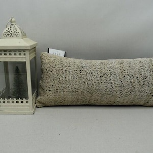May include: A rectangular beige cushion with a textured pattern and a white decorative lantern with a small Christmas tree inside. The lantern has a handle and decorative cutouts.