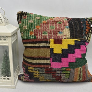 Op de afbeelding: Een patchwork kussen met een verscheidenheid aan kleuren en patronen. Het kussen heeft vierkanten van groene, roze, gele, zwarte en bruine stof. Op de achtergrond staat een witte lantaarn met een kleine kerstboom.