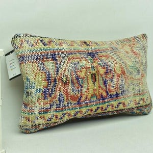 May include: Rectangular cushion with a vintage, patterned design. The cushion features a mix of colours including red, blue, yellow, and green. The cushion has a textured appearance, suggesting a woven or embroidered fabric. A tag is attached to the cushion.