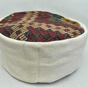 May include: A round, white fabric ottoman with a woven top. The top features a geometric pattern in red, brown, green, and blue. The ottoman is a low, cushioned seat.