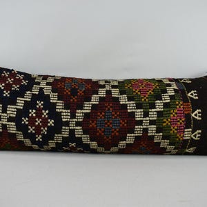 May include: A long, rectangular cushion with a black, white, red, green and blue geometric pattern. The cushion is made of a woven fabric and has a fringe trim.