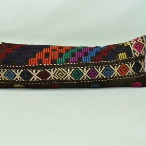 May include: A long, rectangular cushion with a black background and colourful geometric patterns. The cushion is made of woven fabric and has a fringe on the ends.