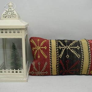 May include: A decorative rectangular pillow with a red, black, and beige geometric pattern. A white metal lantern with a small Christmas tree inside is to the left of the pillow.