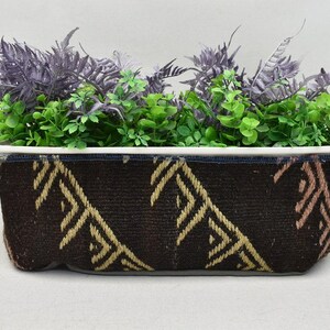 May include: Rectangular planter with a dark brown woven exterior featuring a geometric pattern in tan and pink. The planter is filled with a mix of green and purple faux plants, creating a decorative display.
