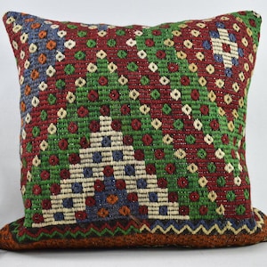 May include: A decorative cushion with a geometric pattern in shades of red, green, blue, and white. The cushion is made of woven fabric and has a fringe border.