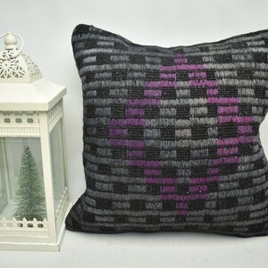 May include: A black and grey patterned cushion with a purple geometric design. The cushion is sitting on a white surface next to a white lantern.