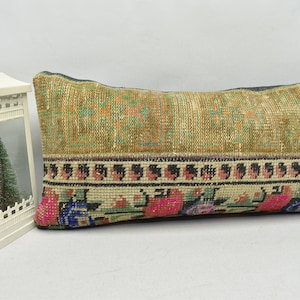 May include: Rectangular decorative pillow with a textured, patterned design. The pillow features a central section in shades of green and orange, with a floral border in pink, blue, and green. The pillow has a vintage, handcrafted aesthetic.