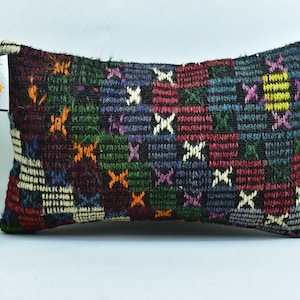 May include: Rectangular pillow with a woven, patchwork design. The pillow features a dark background with squares of red, blue, green, and purple, accented with white cross patterns. A tag is attached to the side.