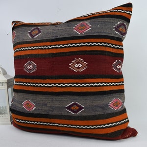 May include: A decorative cushion with a striped pattern in shades of orange, red, black, and grey. The cushion has a geometric design with small diamond shapes.
