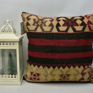 May include: A decorative throw pillow with a woven pattern in shades of red, black, beige, and green. The pillow has a geometric design with intricate details.
