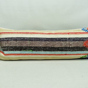 May include: A long, rectangular cushion with a white background and colourful stripes. The cushion has a woven design with geometric patterns in red, blue, green, and black.