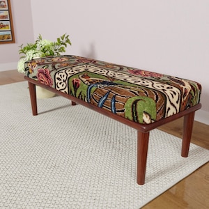 Puede incluir: Un banco rectangular con un coj&iacute;n estampado y patas de madera. El coj&iacute;n presenta un dise&ntilde;o con elementos florales y bot&aacute;nicos en tonos verdes, marrones, rojos y crema. El banco est&aacute; sobre una alfombra clara.