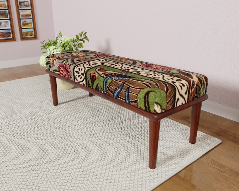 Puede incluir: Un banco rectangular con un coj&iacute;n estampado y patas de madera. El coj&iacute;n presenta un dise&ntilde;o con elementos florales y bot&aacute;nicos en tonos verdes, marrones, rojos y crema. El banco est&aacute; sobre una alfombra clara.