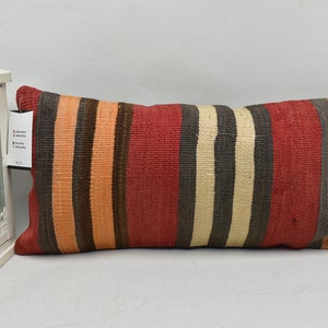 May include: A rectangular cushion featuring vertical stripes in red, orange, beige, and brown. The cushion has a textured finish, possibly woven. A white decorative lantern is visible on the left side.