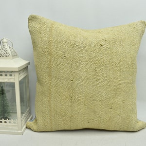 May include: A light beige throw pillow with a woven texture. The pillow is sitting on a white surface next to a white lantern with a small artificial Christmas tree inside.