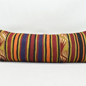 May include: A long, rectangular cushion with a colourful striped pattern in shades of red, orange, yellow, green, blue, and black. The cushion has a geometric design with a repeating pattern of stylised hands.
