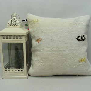 May include: Off-white decorative pillow with embroidered accents in brown, yellow, and orange. A white lantern with a decorative top and a small Christmas tree inside is to the left of the pillow.