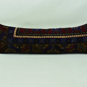 May include: A long, rectangular cushion with a colourful, geometric pattern in shades of red, blue, gold, and brown. The cushion is made of a woven fabric with a textured surface.