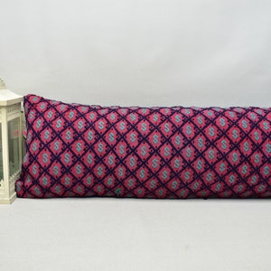 May include: A long, rectangular decorative cushion with a geometric pattern. The cushion features a repeating diamond pattern in shades of pink, teal, and dark blue. The cushion is approximately 76 cm long.