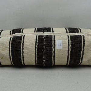May include: Rectangular, striped floor cushion in cream and dark brown. The cushion has vertical stripes and a zipper closure. A small white tag with text is attached to the cushion.