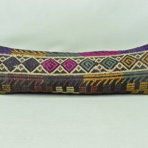 May include: A long, rectangular cushion with a woven, geometric pattern. The cushion features a dark background with colourful diamond and zigzag designs in shades of pink, yellow, green, and purple. The cushion is likely for home decor.