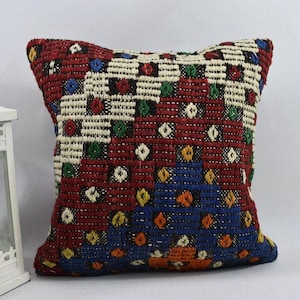 May include: Square cushion with a geometric pattern. The cushion has a red background with a woven design in white, blue, green, and orange. The cushion is square-shaped.