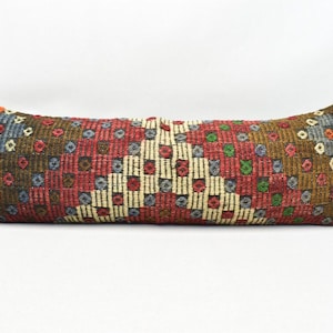May include: A long, rectangular cushion with a colourful geometric pattern. The cushion is made of woven fabric and has a brown, red, white, blue, and green colour scheme.
