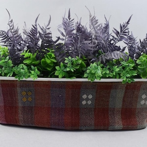 May include: A rectangular planter with a plaid pattern in shades of red, brown, and gray. The planter is filled with artificial plants, including ferns and green foliage.