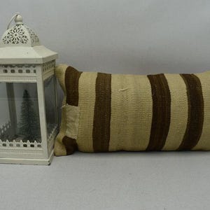 May include: A rectangular cushion with vertical brown stripes on a beige background. A white metal lantern with a decorative top and glass panels is to the left of the cushion. The lantern has a small Christmas tree inside.