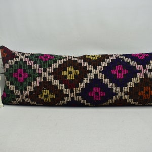 May include: A long, rectangular pillow with a colorful geometric pattern. The pattern is made up of squares and diamonds in shades of pink, green, brown, yellow, and blue. The pillow is made of a soft, woven fabric.